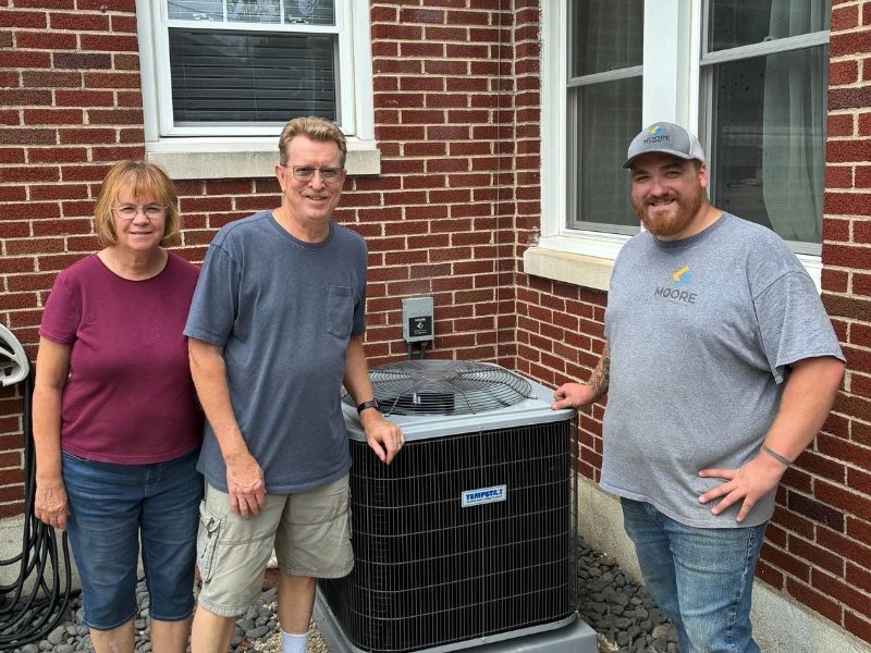 Moore Comfort Heating & Cooling team member standing with two homeowners beside a residential outdoor air conditioning unit