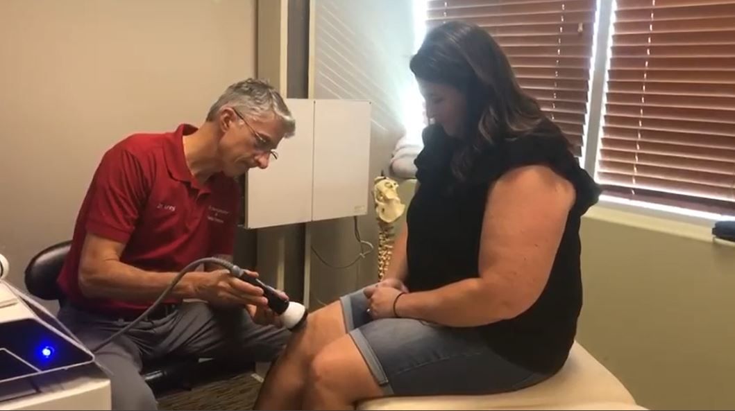 Man using medical device on woman's knee in a doctor's office. She sits, looking down.