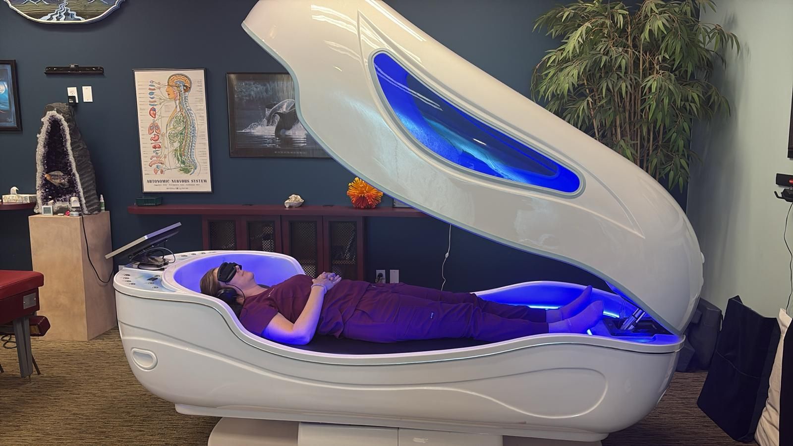 A woman lies in a white spa pod with blue interior lights. The pod is in a room with decor.