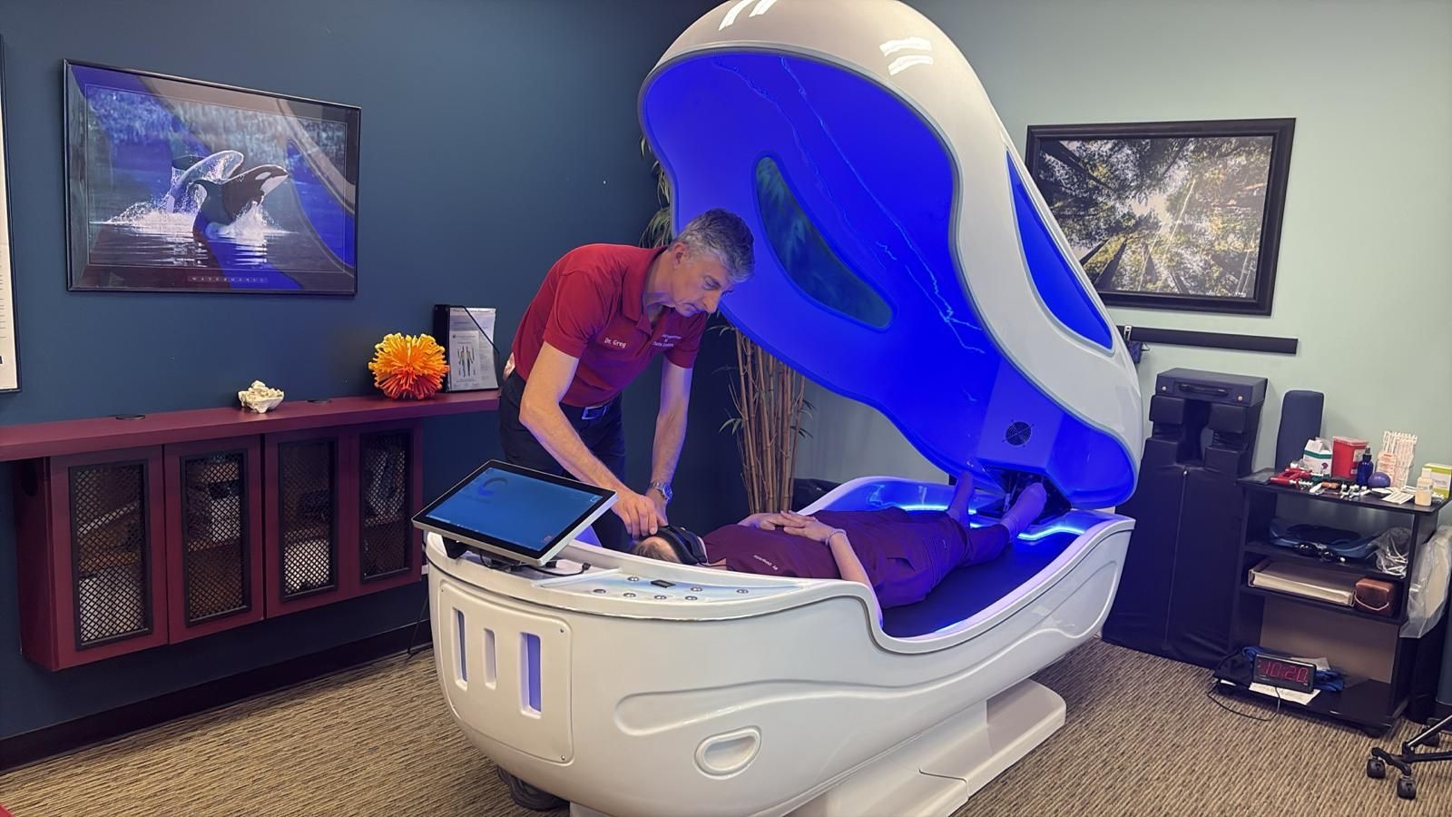 Dr Greg adjusting controls on a white wellness pod with a person inside. Blue light glows from the pod.