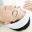 Person with a white headband, eyes closed, having a facial treatment in a spa setting.