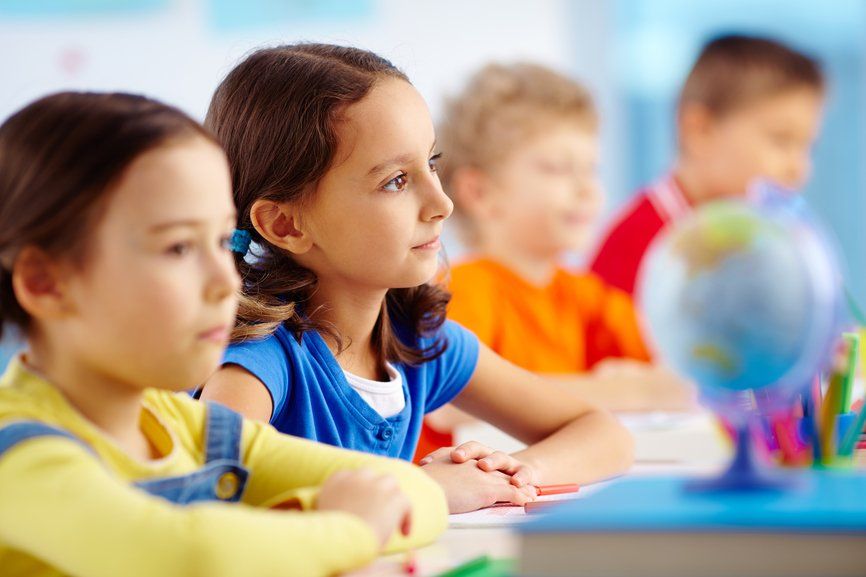 Children in a classroom listening attentively; a globe and supplies are on the desk.