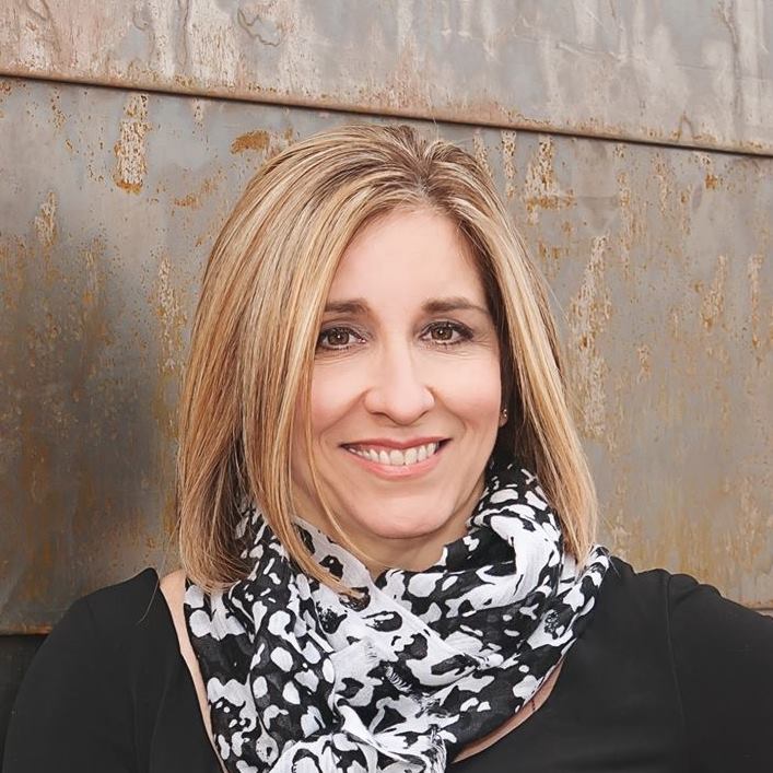 Woman with blonde hair smiles, wearing a black shirt and patterned scarf, stands in front of a rusty metal wall.