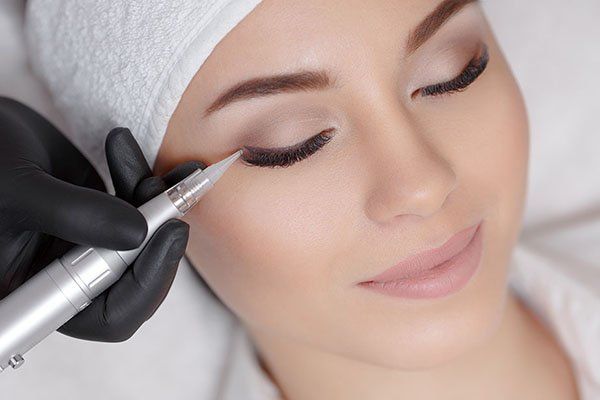 A woman undergoing eyeliner permanent makeup; technician's gloved hand holds the device near the eye.