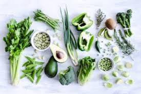Variety of green vegetables and herbs on a white surface; including avocado, peppers, and broccoli.