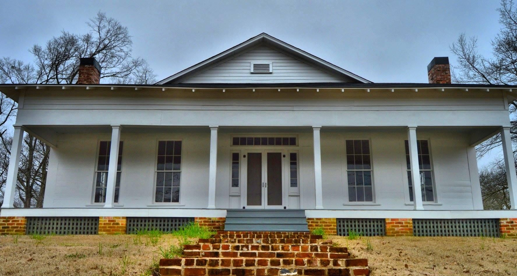 White farmhouse with wraparound porch and brick steps under a cloudy sky