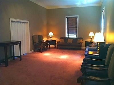 Empty waiting room with red carpet, chairs along the wall, and two lit lamps near a sofa.
