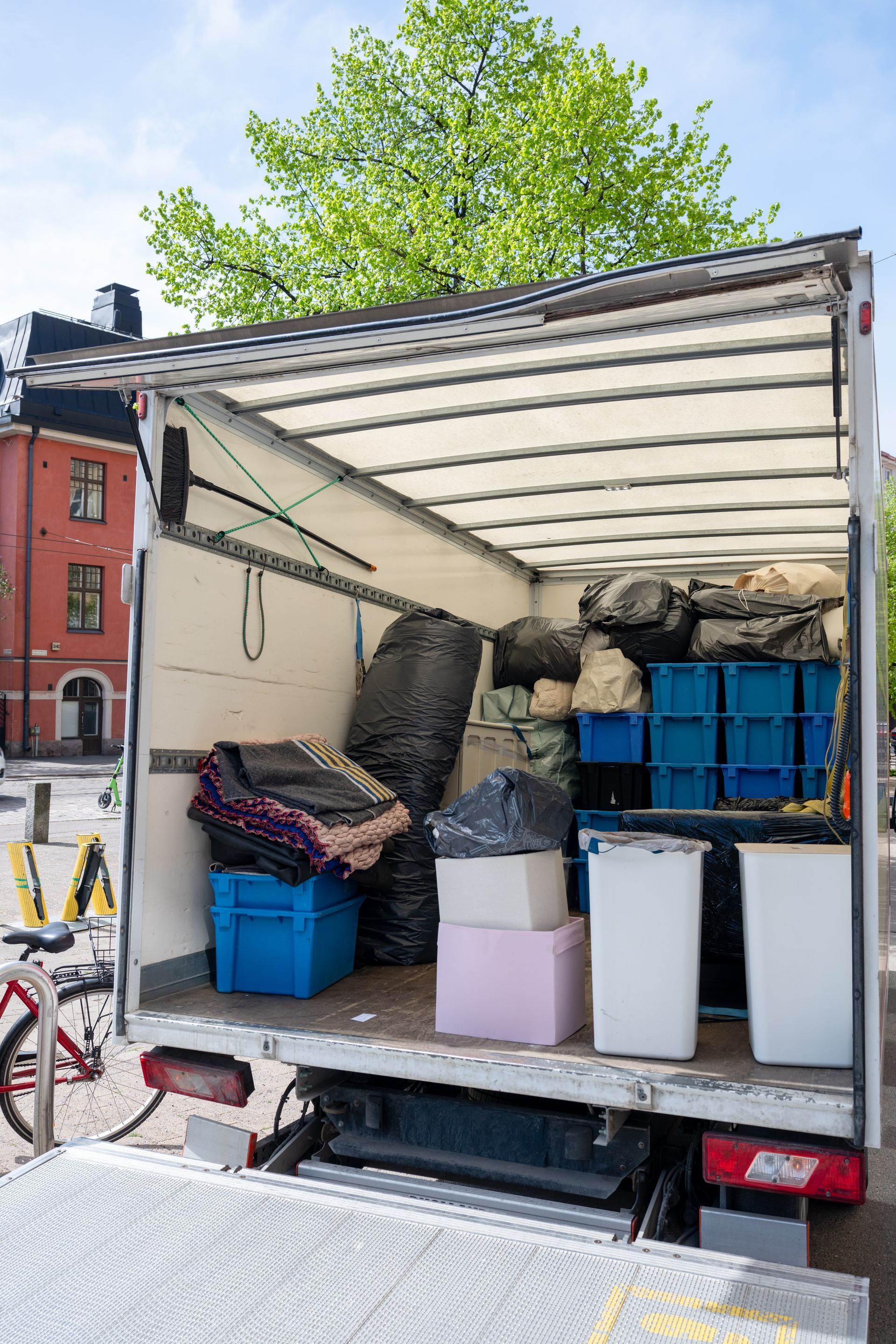 Truck with open cargo area loaded with bins, bags, and boxes at a street curb.
