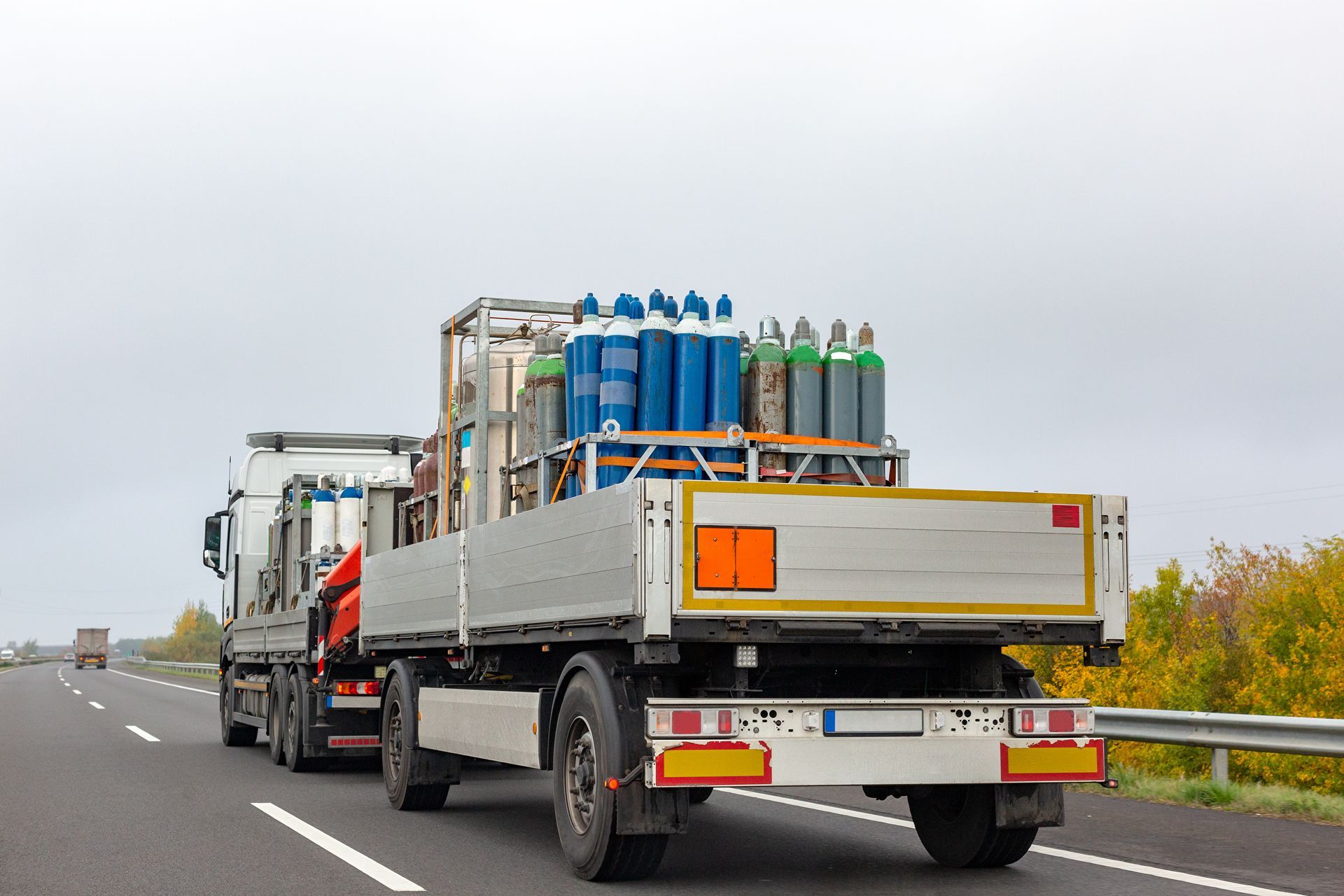 Flatbed truck carrying stacked gas cylinders driving on a wet highway under gray skies