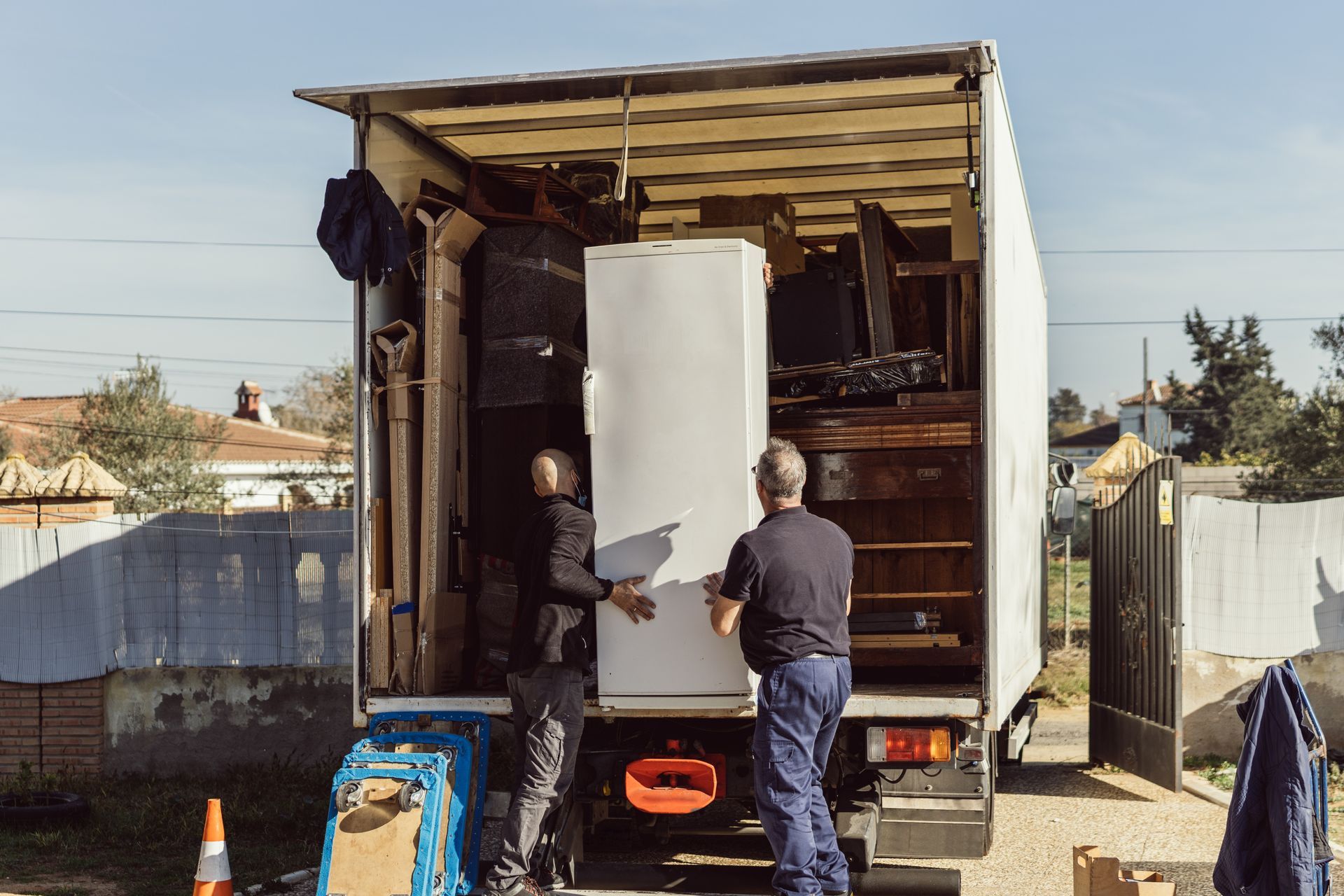 Two workers loading a white refrigerator into a truck in a yard, with tools and equipment nearby.