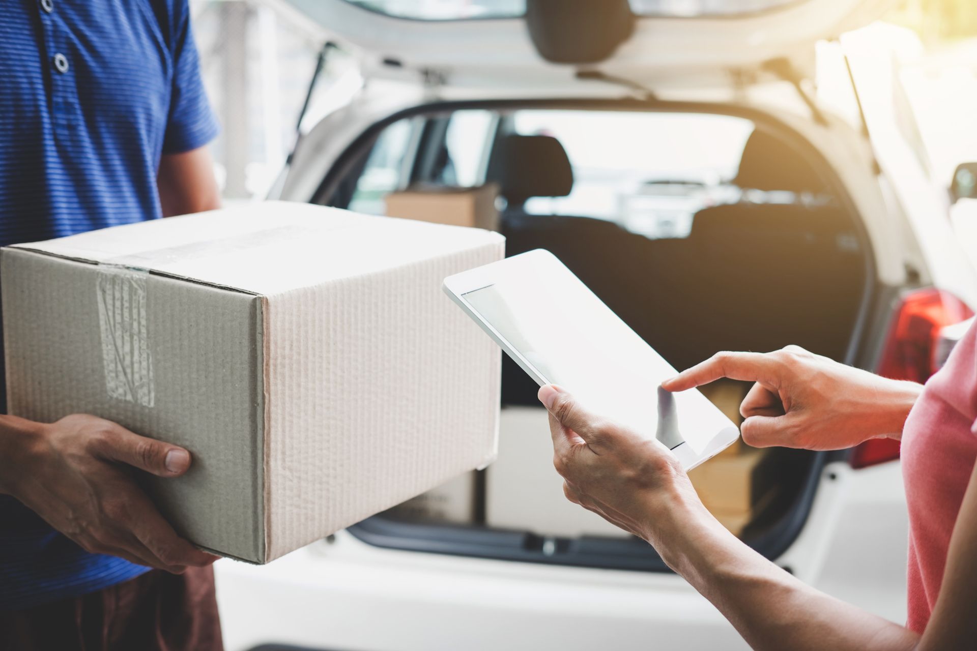 A delivery worker holds a cardboard box while a customer signs for it on a tablet near the open trunk of a car.