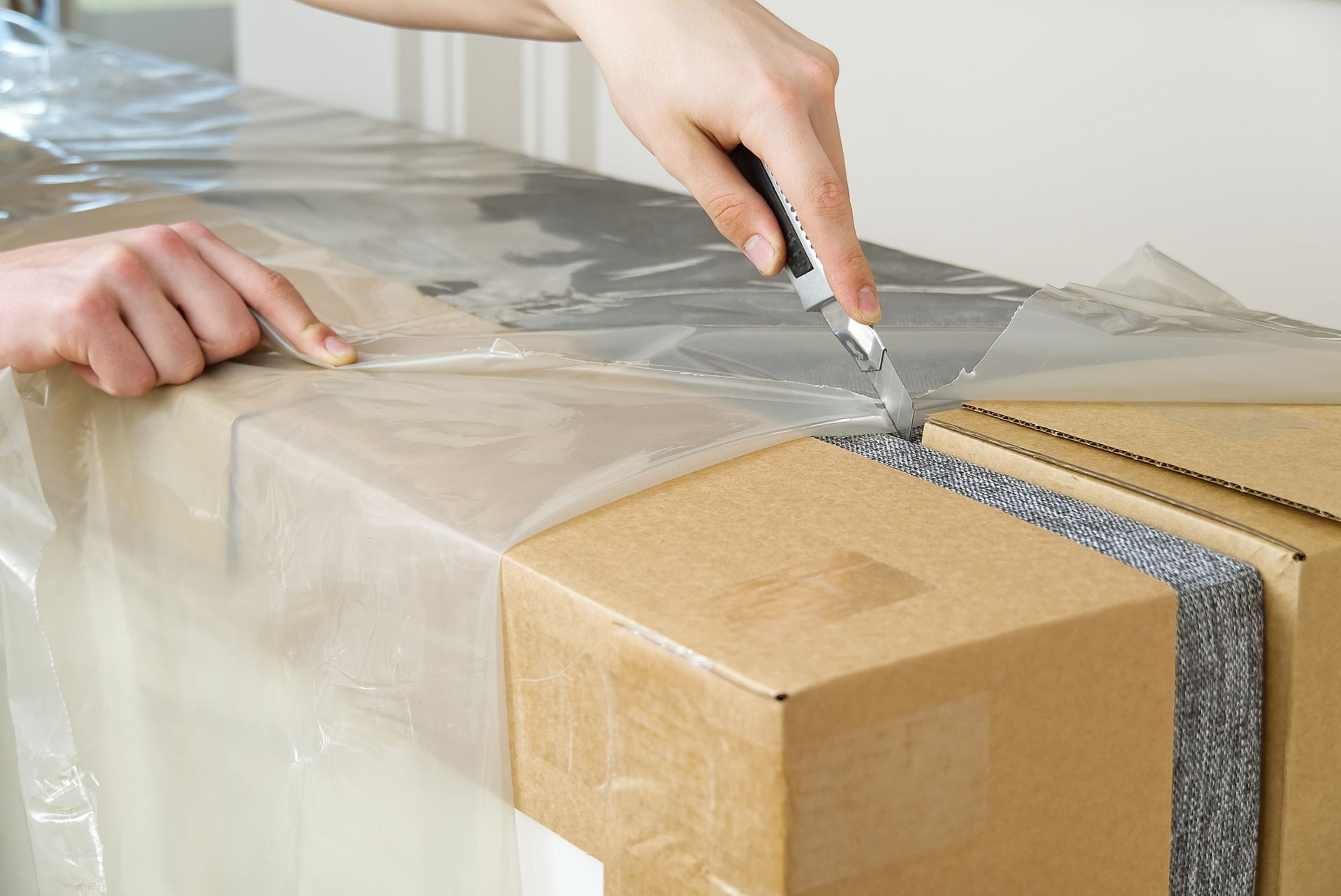 Hands use a utility knife to cut away clear plastic protective film from a cardboard package.