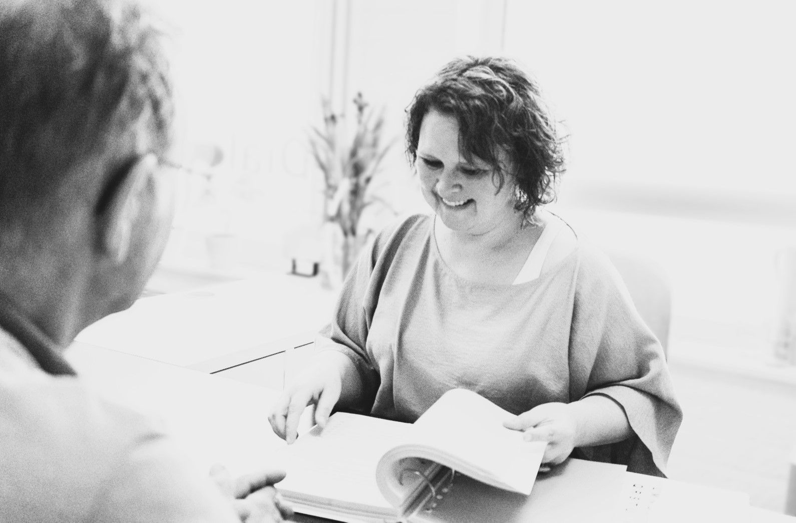 Een vrouw zit aan een tafel en leest een boek voor aan een man.
