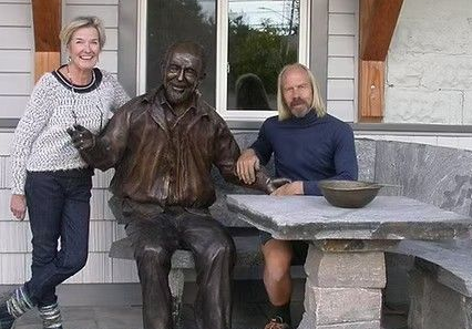 Woman standing beside bronze statue of seated man at outdoor stone table on a porch
