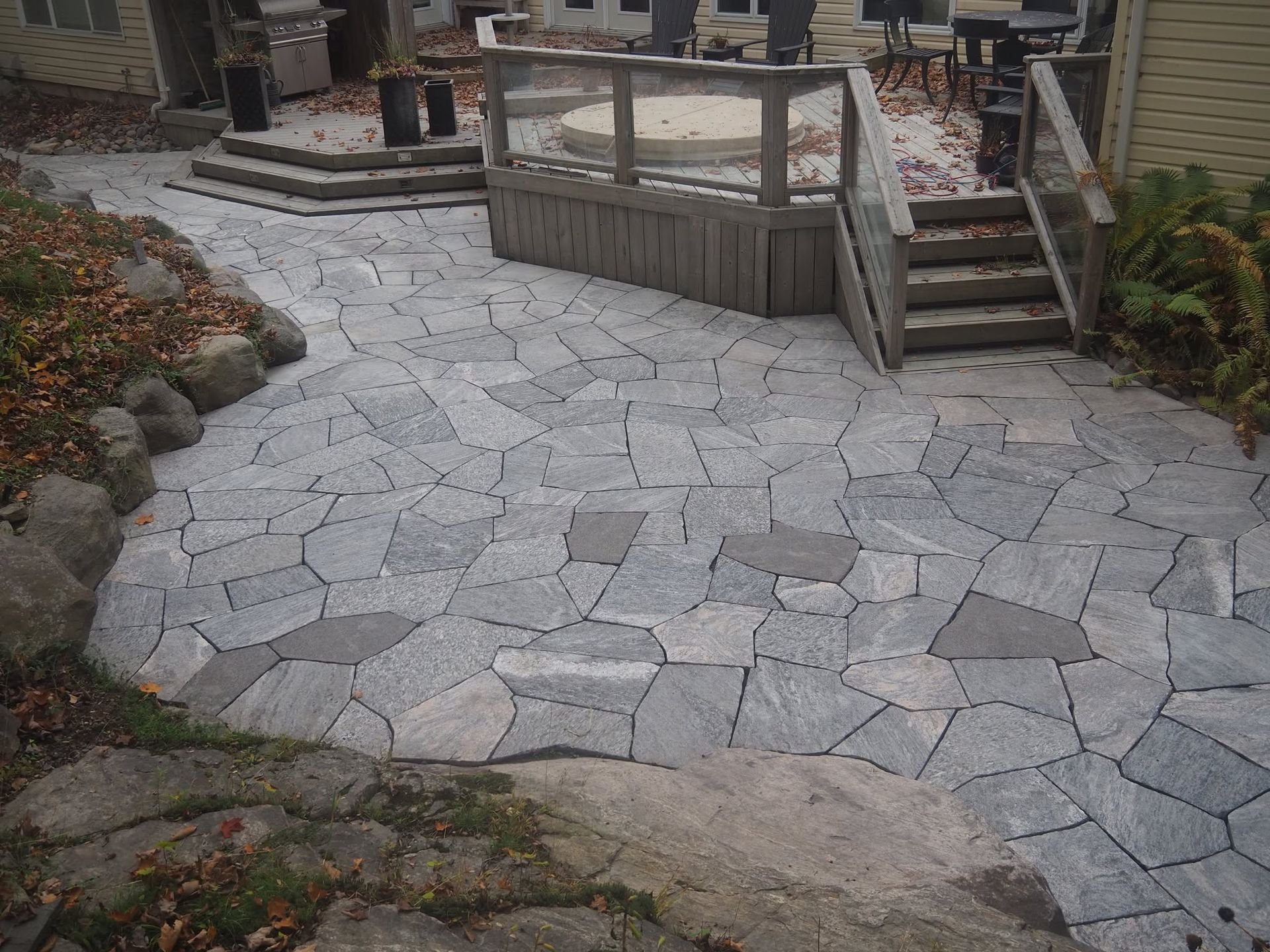 Stone patio with irregular gray pavers and a small wooden deck and stairs in a garden setting