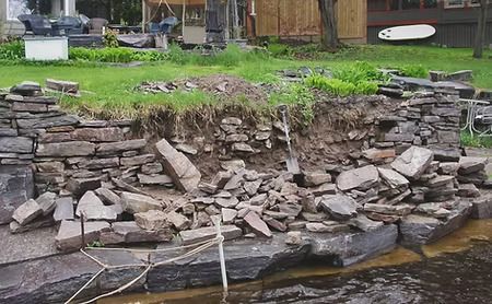 Collapsed stone retaining wall by a grassy yard, with rubble scattered along the base and water below