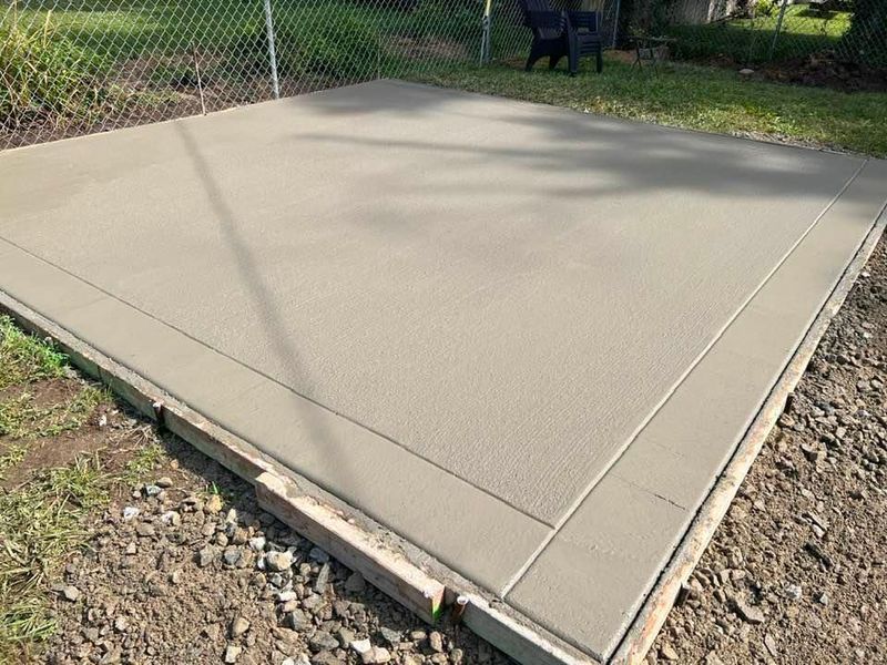 A newly poured concrete pad, light gray in color, framed by wooden borders, set in a grassy yard.