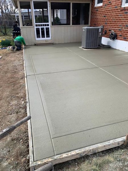 Newly poured concrete patio next to a brick house; a worker is smoothing the surface.