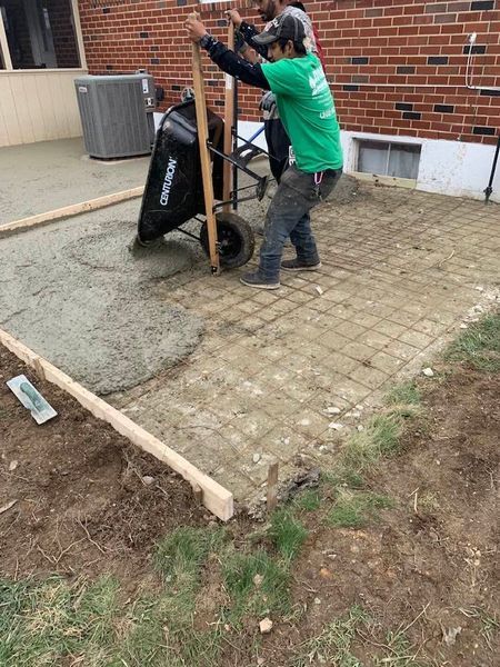 Construction worker pours concrete from a wheelbarrow onto rebar, building a patio. Outdoors, next to a building.