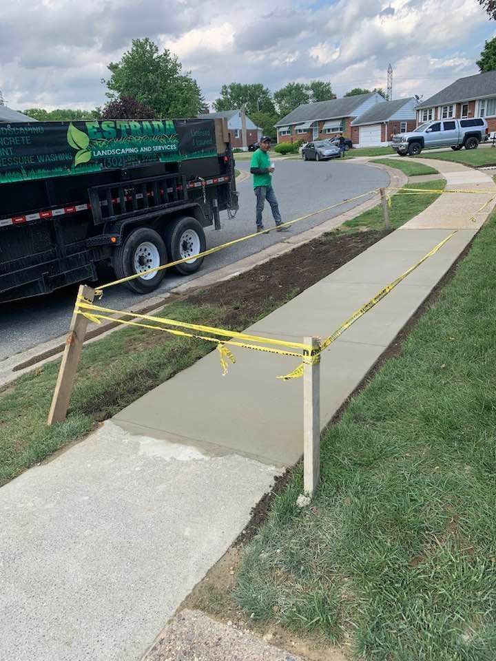 New concrete sidewalk being poured, with a landscaping truck and worker present. Yellow caution tape.