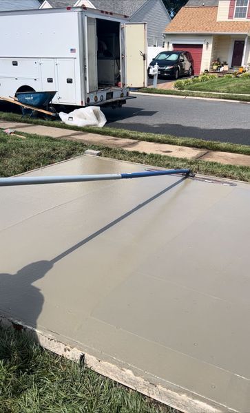 Freshly poured concrete driveway being smoothed with a long tool; a work truck is parked nearby.