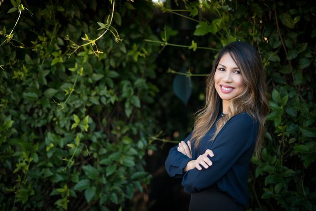 A woman is standing with her arms crossed in front of a bush.