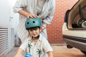 A woman is putting a helmet on a little boy who is riding a scooter.