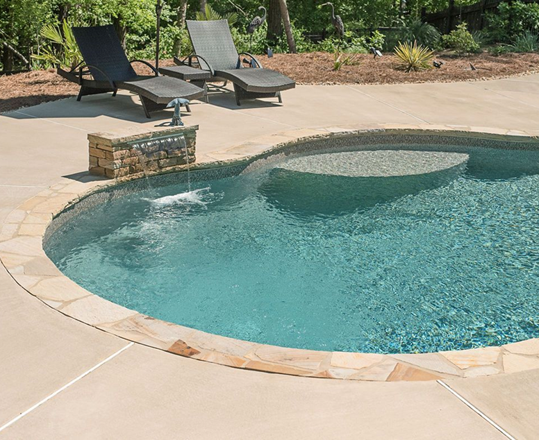 Swimming pool surrounded by stone patio, deck, and trees; calm blue water.