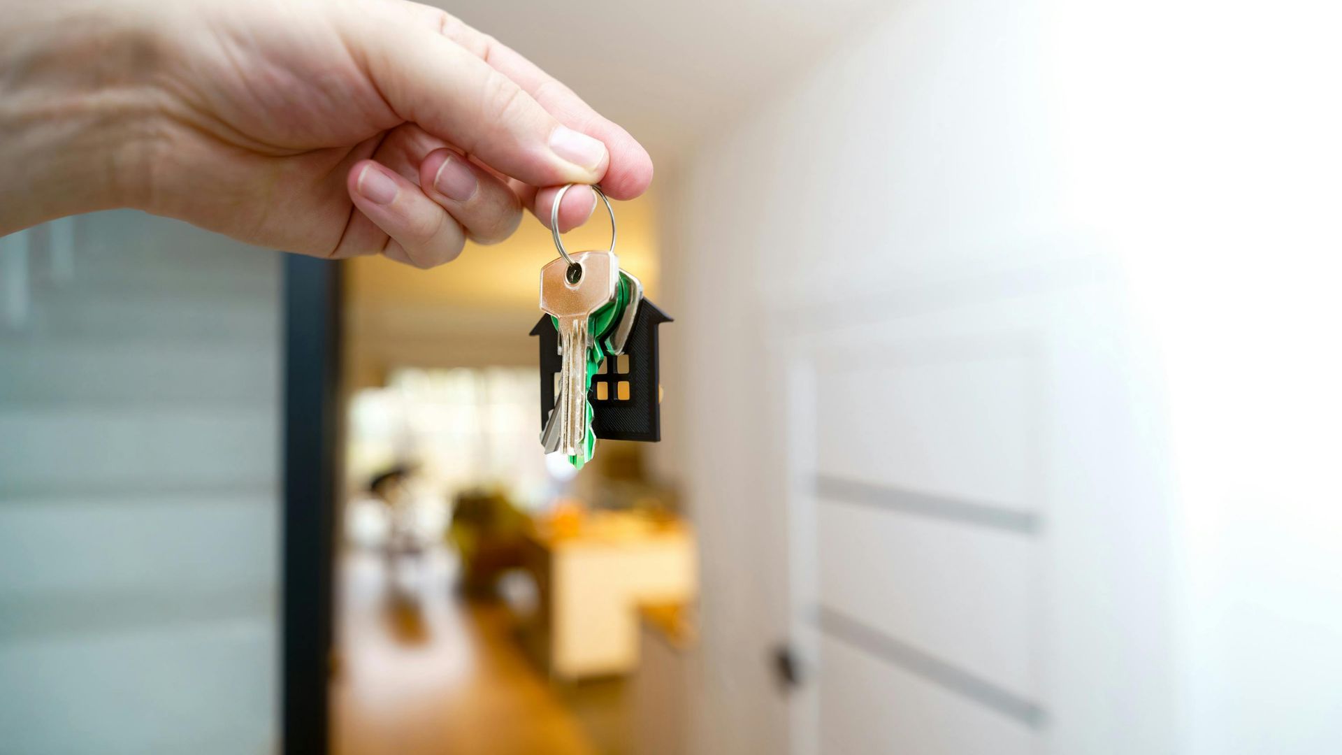 Two people sign an insurance document on a desk featuring a small house model, keys, cash, and a calculator.
