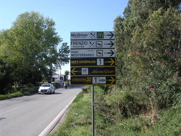 Cartelli stradali a lato di una strada rurale con alberi e un'auto bianca di passaggio
