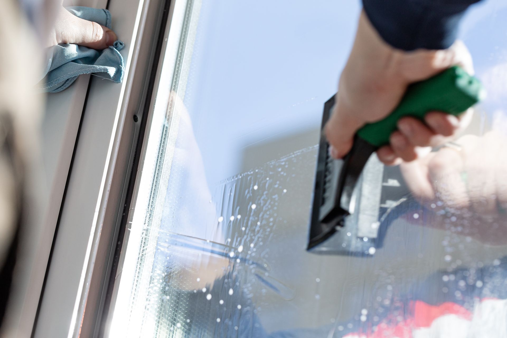 A hand uses a squeegee to clean a glass window, with another hand holding a cloth to wipe the frame.