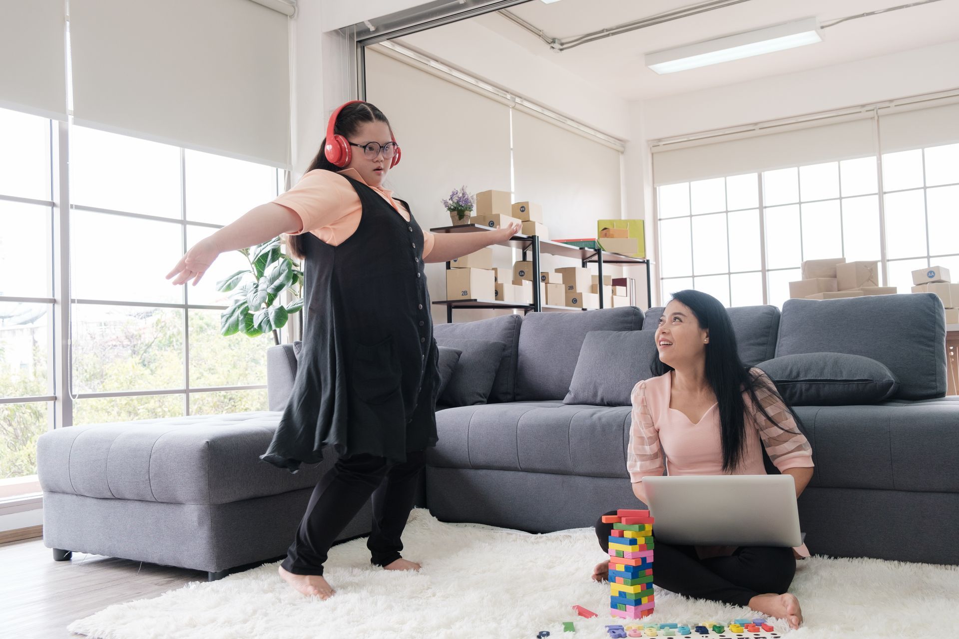 Person wearing headphones dancing while another sits with a laptop and colorful blocks on floor.