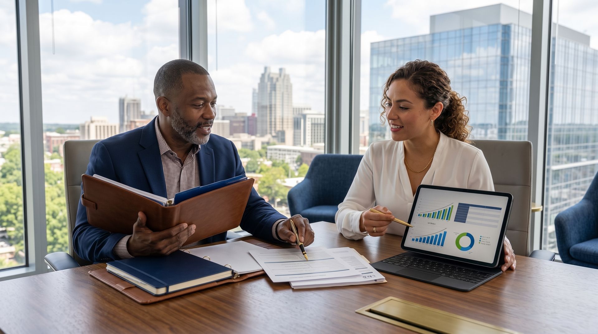 Two coworkers review documents at a conference table with a laptop showing charts, city skyline behind them.