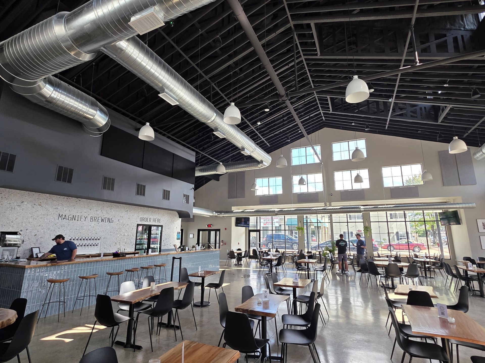 Interior of a spacious brewery with a bar, tables, chairs, and large windows. Someone stands behind the bar.
