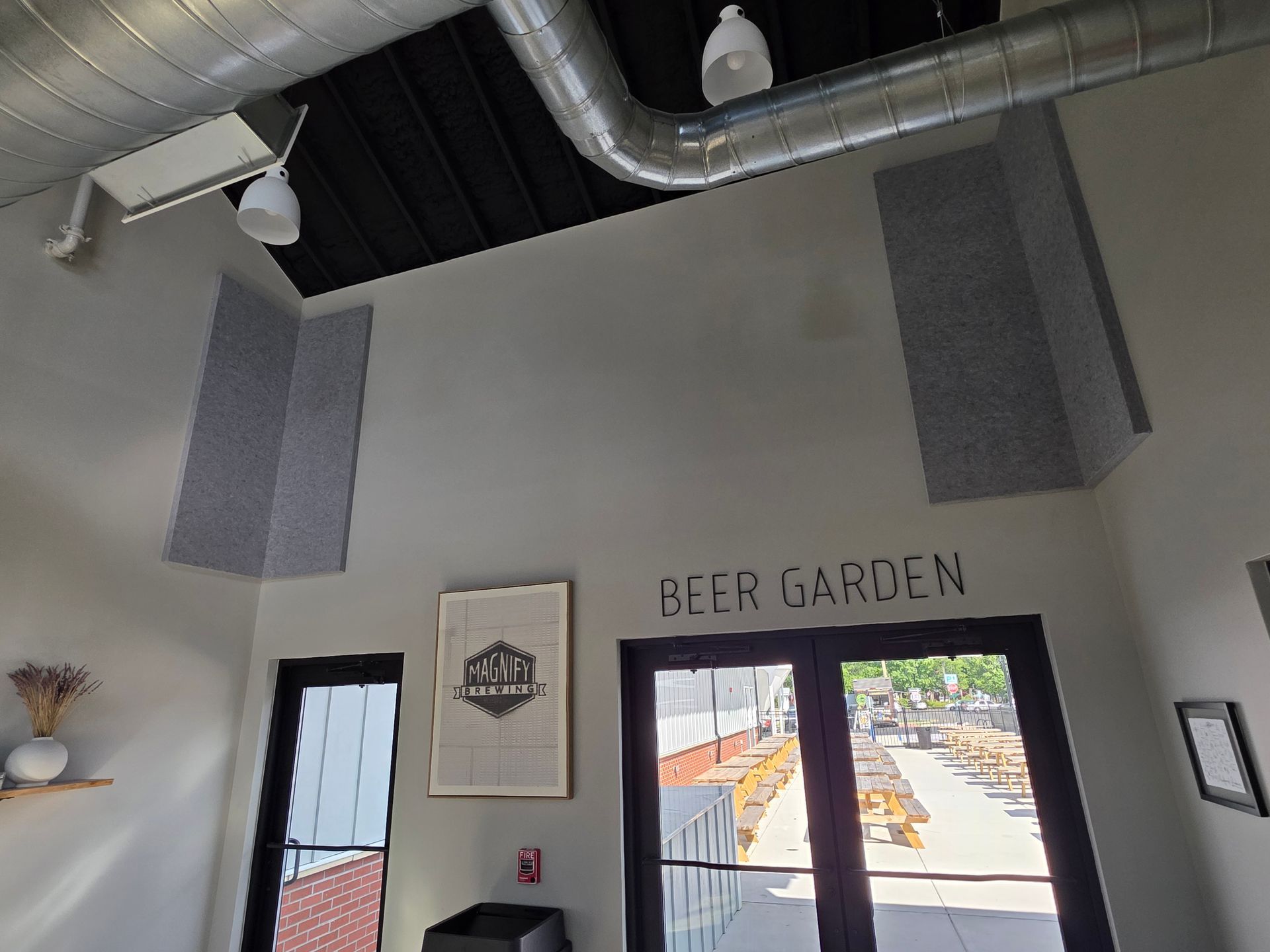 Interior of a beer garden with gray acoustic panels, ductwork, and outdoor seating visible through doors.