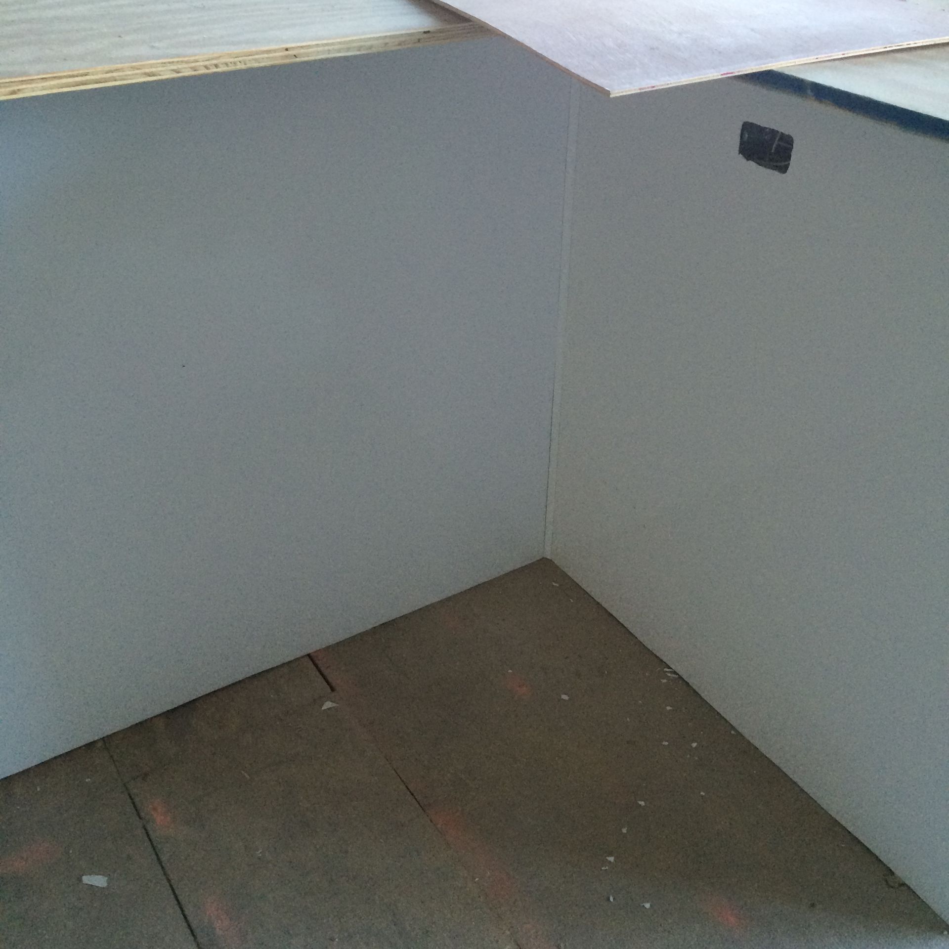 White painted corner of a built-in shelf on a wooden floor, partially covered by light-colored plywood.