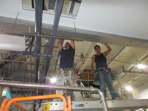 Two workers installing metal supports in a high-ceilinged industrial space. One holds a support bar, other steadies.