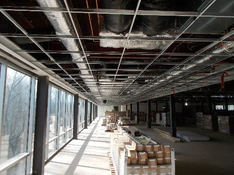 Interior construction site: metal grid ceiling, ductwork, windows, materials in a long, sunlit corridor.