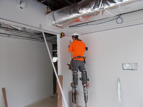 Construction worker on stilts using a laser level in a room with white walls, unfinished ceiling.