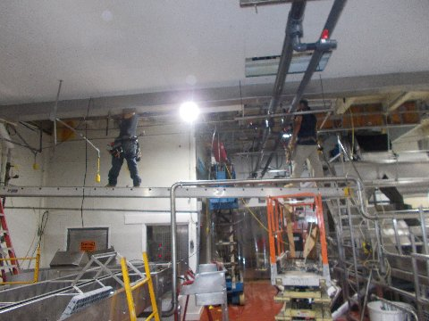 Workers on scaffolding installing equipment in a factory setting. Metallic pipes and machinery are visible.
