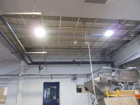 Industrial interior with exposed ceiling grid, pipes, wiring, lights, and equipment. A blue door is visible.
