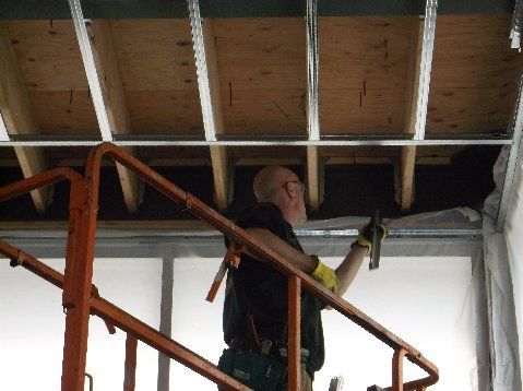 Person on an orange lift works on a ceiling, holding a tool. Wooden beams and metal framing are visible.