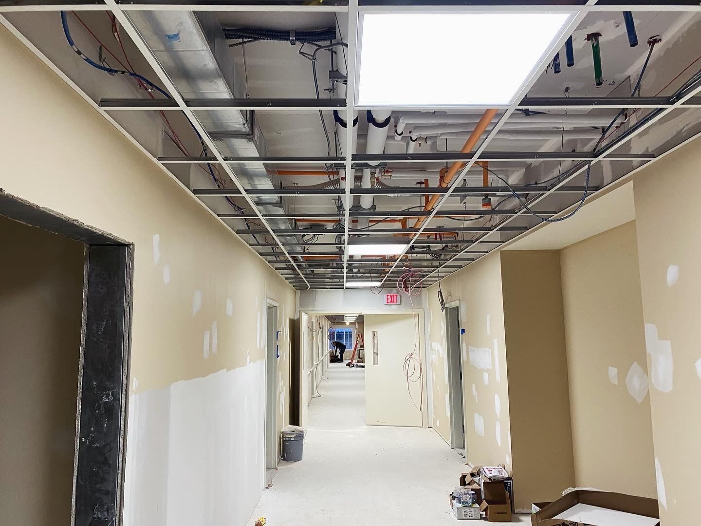 Hallway under construction, exposed ceiling grid, beige walls, doorways, overhead lighting.