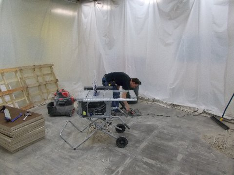 Person using a table saw in a room with plastic sheeting on the walls.  Wood and tools are nearby.