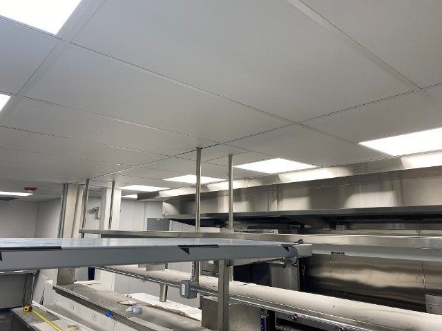 Stainless steel kitchen with suspended ceiling, overhead shelving, and multiple bright lights.