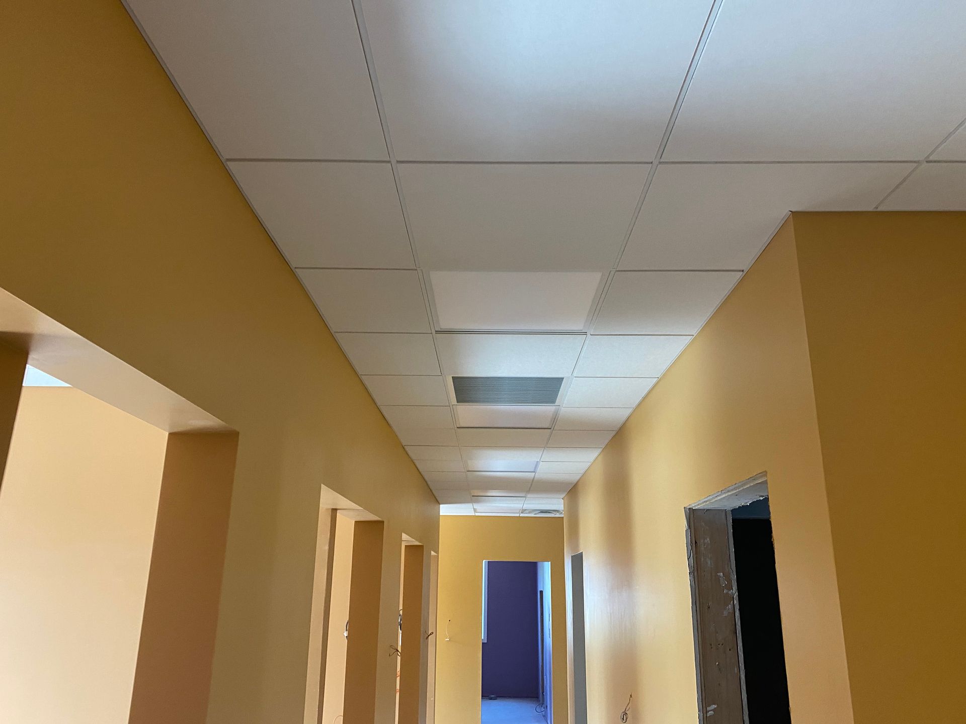 Narrow hallway with yellow walls, white ceiling tiles, and open doorways.