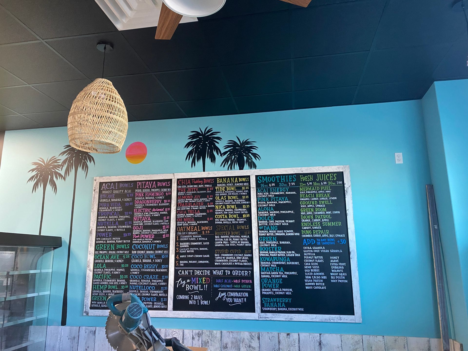 Restaurant interior: blackboard menu, turquoise wall, palm tree silhouettes, wicker pendant light, white counter.