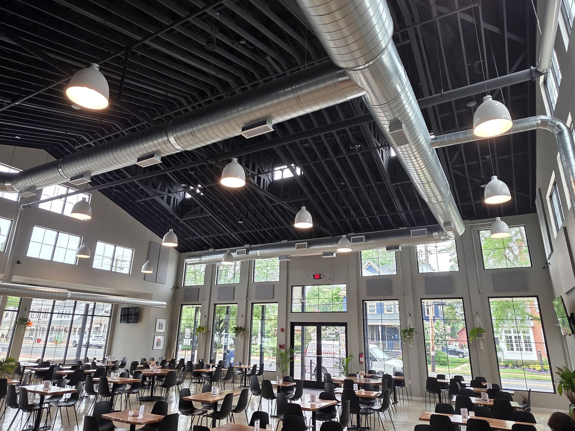 Interior of a restaurant with black ceiling, ductwork, pendant lights, tables, and large windows.