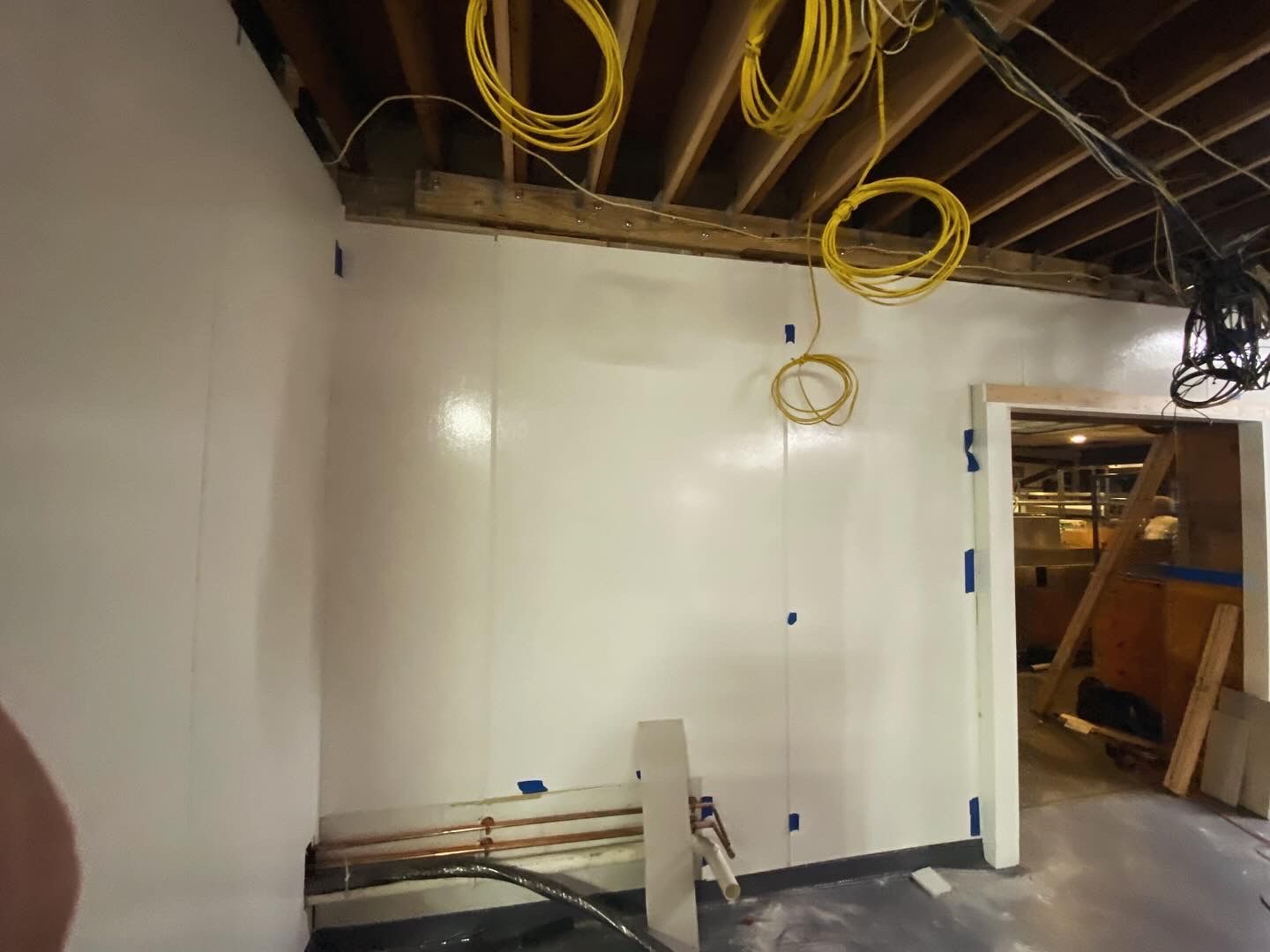 Interior view of a room under construction with white panel walls and exposed ceiling beams. Electrical wires hang.