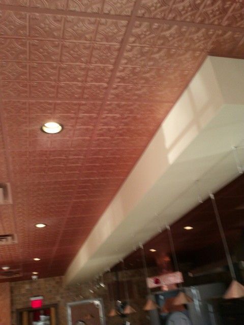 Copper-colored, patterned ceiling with recessed lights, and a white, rectangular structure in a dining area.